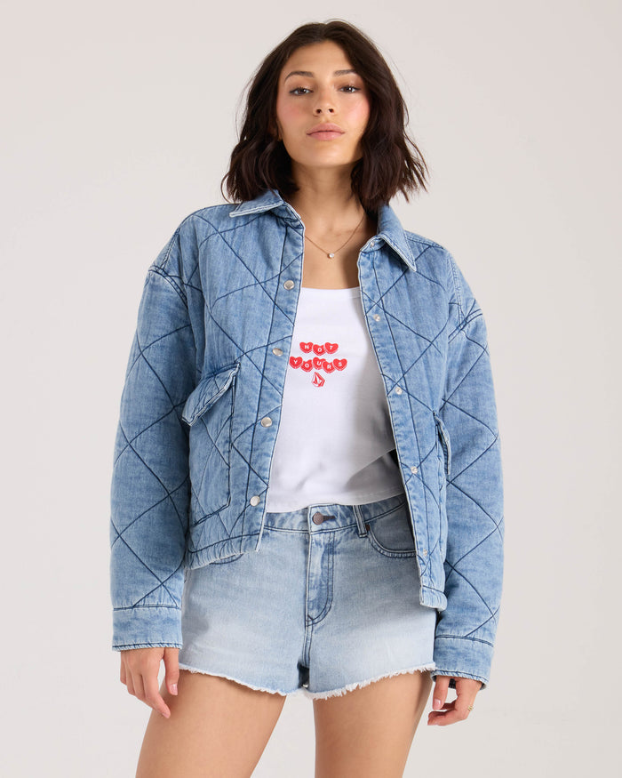 A woman wears the Volcom Women's OC Ramps quilted light blue denim jacket with a boxy fit over a white top with red text and light denim shorts, standing against a plain light background.