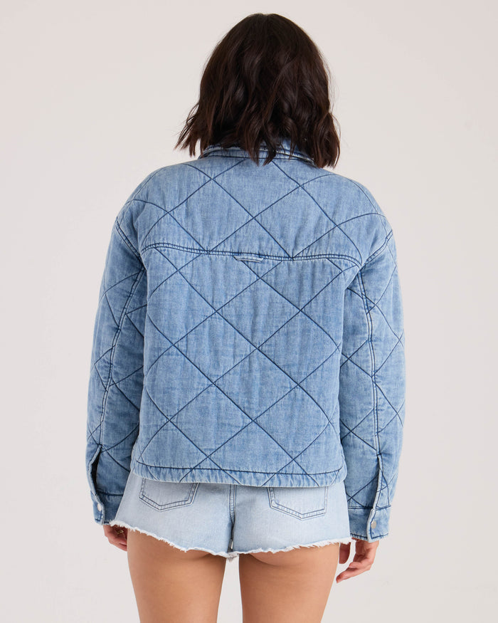 A woman with shoulder-length dark hair is seen from the back, wearing Volcom's Women's OC Ramps light blue boxy denim jacket and light denim cut-off shorts against a plain background.