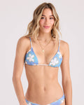 A woman with long, wavy blonde hair models the Volcom Women's Ocean Haze Triangle Top, a blue floral bikini with thin straps. She stands against a plain light background, smiling softly at the camera and wearing gold layered necklaces.