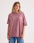 A woman wearing the Volcom Women's Off The Grid Bad Tee—an oversized mauve graphic T-shirt made from 100% cotton with white text on the chest—paired with blue jeans and standing against a plain light background.