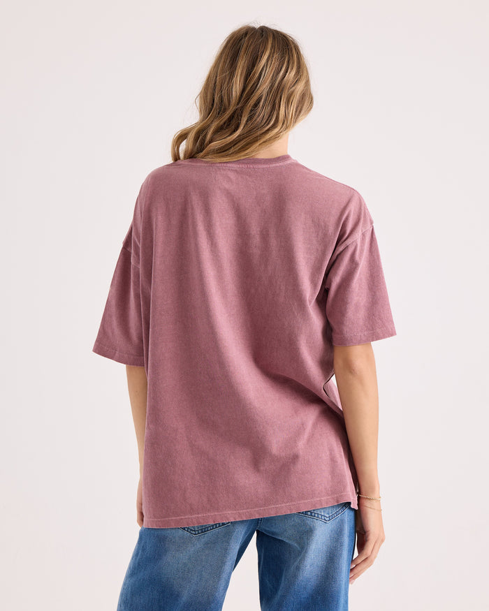 A person with long, wavy hair is seen from behind wearing the Volcom Women's Off The Grid Bad Tee, an oversized mauve 100% cotton t-shirt, paired with blue jeans and standing against a plain light background.