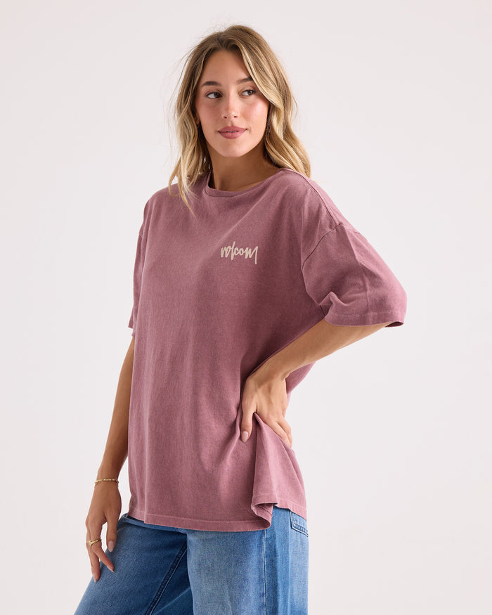 A woman models the Volcom Women's Off The Grid Bad Tee, an oversized mauve graphic t-shirt with "bloom" across the front, paired with blue jeans against a plain light backdrop. Made from 100% cotton, this tee combines comfort and effortless style.