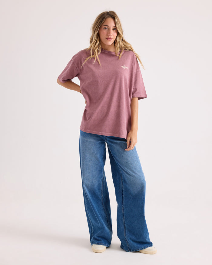 A person stands against a plain background wearing the Volcom Women's Off The Grid Bad Tee, an oversized mauve t-shirt made from 100% cotton, paired with wide-leg blue jeans and white shoes. They have long, wavy hair and strike a relaxed pose with one hand by their side and the other on their hip.