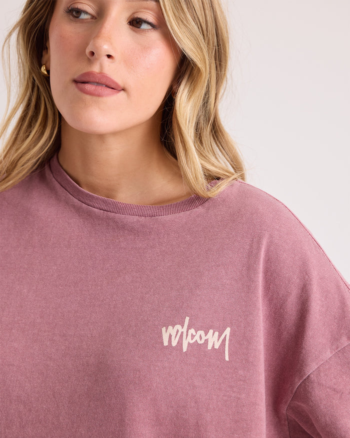A woman with wavy blonde hair wears the Volcom Women's Off The Grid Bad Tee in mauve and looks slightly to the side with a neutral expression. The background is plain and light-colored.