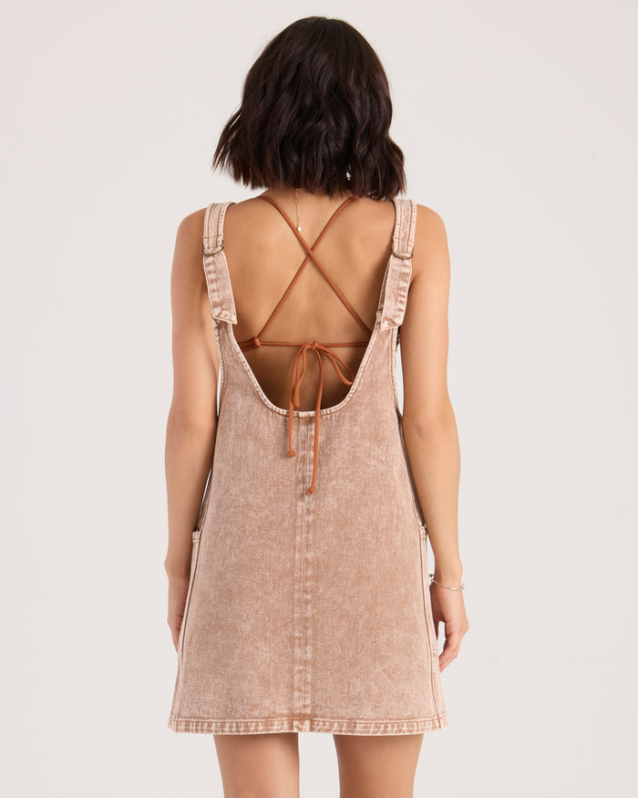 A woman with shoulder-length dark hair is seen from the back, wearing the Volcom Women's Ollie light brown A-line denim skirtall with wide straps and a criss-cross tie open back. She stands against a plain, sun-warmed henna-toned background.