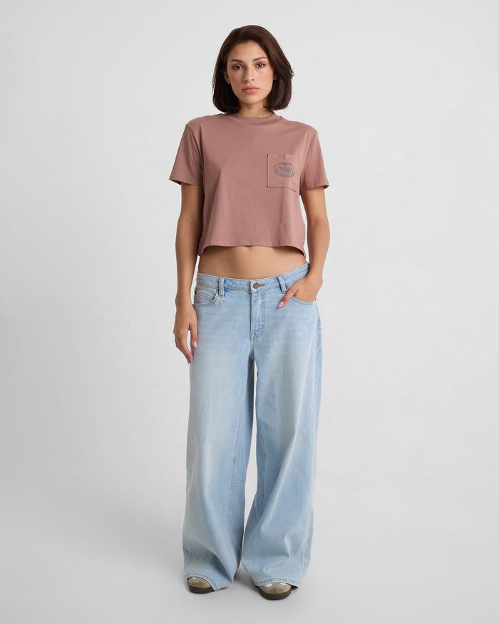 A woman stands against a white background wearing the Volcom Women's Pocket Dial Short Sleeve Tee and loose, light blue wide-leg jeans, with one hand in her pocket and a neutral expression.