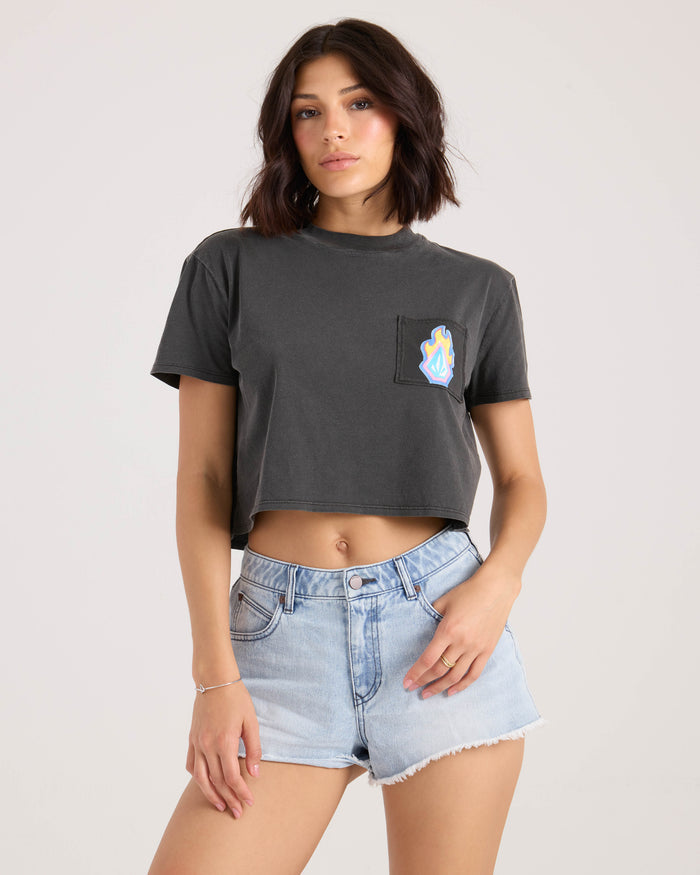 A woman with shoulder-length dark hair wears the Volcom Women's Pocket Dial Tee, a vintage black cropped crew neck with a colorful patch pocket, paired with light blue denim shorts. She stands against a plain, light background.