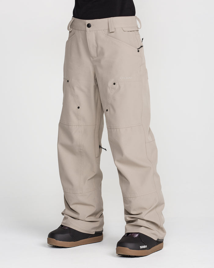 A person wearing Volcom Women’s Pollock Baggy Pants in light beige, featuring multiple pockets and visible seams, paired with black snow boots. The loose-fitting pants are shown from the waist down against a plain white background.