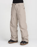 A person wearing Volcom Women’s Pollock Baggy Pants in light beige, featuring multiple pockets and visible seams, paired with black snow boots. The loose-fitting pants are shown from the waist down against a plain white background.