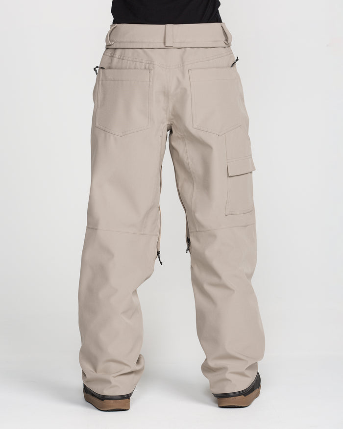 A person is shown from the back, standing on a white background, wearing Volcom Women’s Pollock Baggy Pants in beige. The pants feature multiple pockets, and brown-soled shoes are visible.