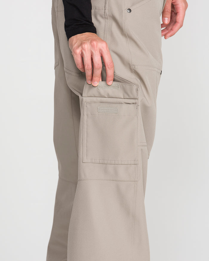 A person wearing the Volcom Women’s Pollock Baggy Pants—beige, with visible stitching and functional pockets reminiscent of snow pants—rests their hand on a side pocket. The background is plain and light-colored.