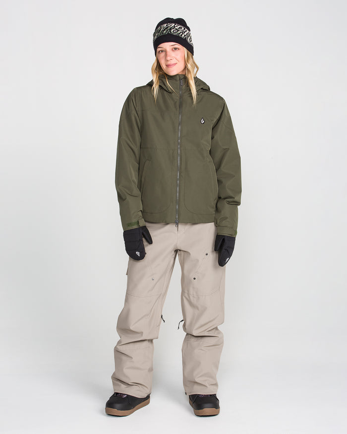 A person wearing an olive green jacket, Volcom Women’s Pollock Baggy Pants in beige, black gloves, black boots, and a black beanie stands and smiles against a plain white background.