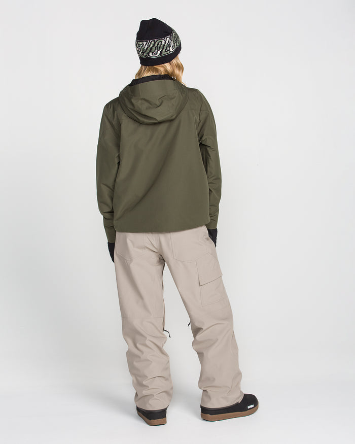 A person stands facing away, dressed in a dark green jacket, black gloves, boots, and a black beanie with white writing, paired with Volcom Women’s Pollock Baggy Pants in light beige. The background is plain white.