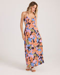 A woman with light brown hair wears the Volcom Women's Post Punk Dress, an A-line maxi with a bold floral print in orange, purple, pink, and green. She pairs it with sandals and stands against a plain white background.