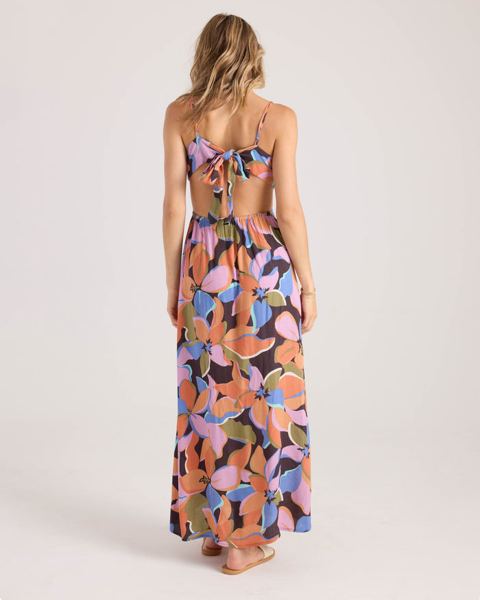 A woman stands barefoot facing away, wearing the Volcom Women's Post Punk Dress—an A-line maxi featuring a colorful floral and abstract leaf print, thin straps, and an open back with a large bow detail.