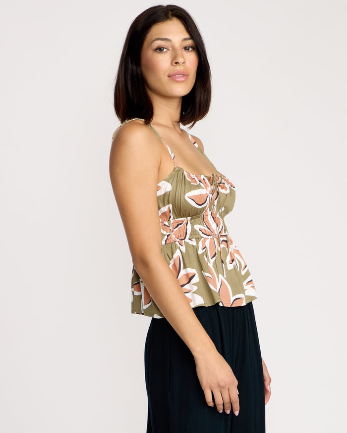 A woman with straight dark hair models the Volcom Women's Psychonic Cami—an olive green matte satin top with pink and white florals and adjustable straps—paired with a dark skirt, standing sideways against a plain light background.