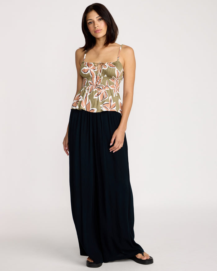 A woman faces forward in a Volcom Women's Psychonic Cami featuring adjustable straps and a smocked bodice, paired with a loose black ankle-length skirt and black sandals, against a plain white background.
