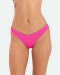 A person models Volcom’s Women's Pucker Up 90S V Bikini Bottom in bright pink textured recycled fabric, shown from mid-torso to upper thighs against a plain white background with a small butterfly tattoo on the left torso.