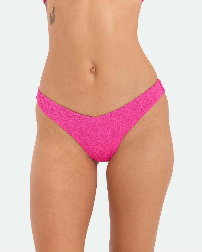 A person models Volcom’s Women's Pucker Up 90S V Bikini Bottom in bright pink textured recycled fabric, shown from mid-torso to upper thighs against a plain white background with a small butterfly tattoo on the left torso.