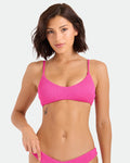 A woman with short brown hair wears the Volcom Women's Pucker Up Bralette in fuchsia, featuring adjustable straps. She stands against a plain light background, looking at the camera with a neutral expression and a small butterfly tattoo on her left ribcage.
