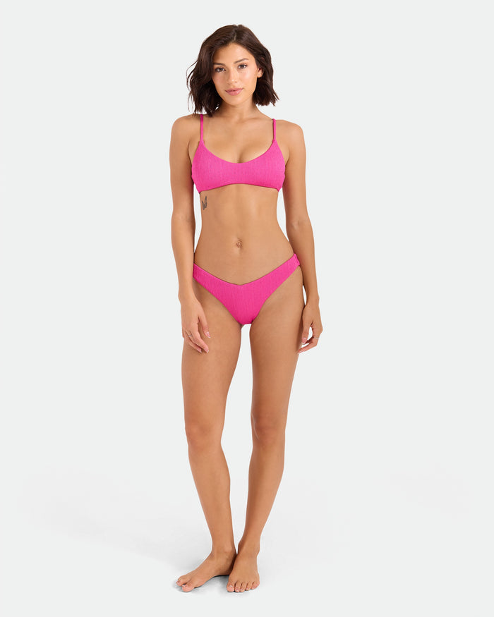 A woman with short brown hair stands barefoot against a white background, wearing the Volcom Women's Pucker Up Bralette in fuchsia. The bikini features adjustable straps and is made from recycled stretch fabric. She faces forward with a neutral expression.