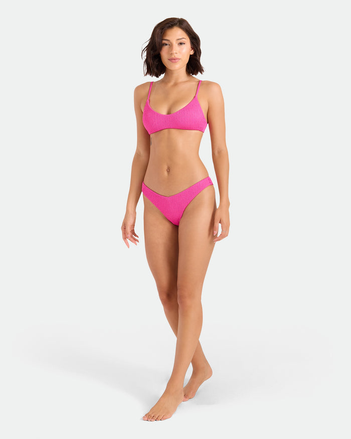 A woman with shoulder-length brown hair walks barefoot against a plain white background, wearing Volcom's Women's Pucker Up Bralette, a bright pink recycled stretch bikini top with adjustable straps.