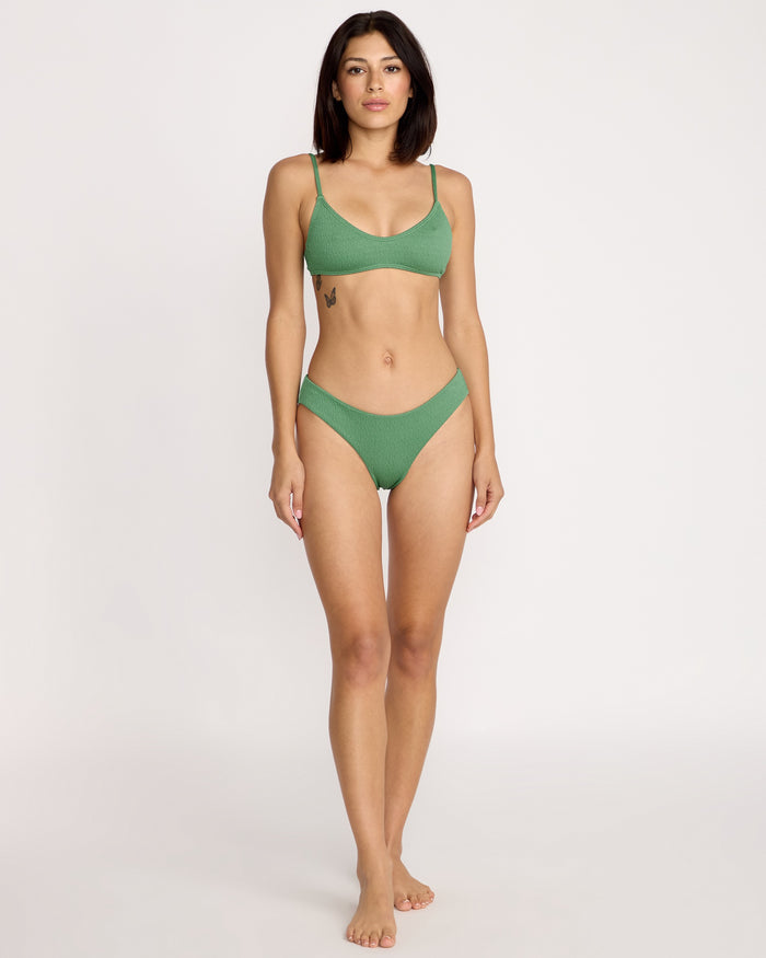 A woman stands barefoot against a light background, wearing the Volcom Women's Pucker Up Scoop Crop Bikini Top in green with thin straps and textured recycled nylon fabric. She has straight dark hair, a neutral expression, and a left ribcage tattoo.