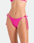 A model wears the Volcom Women's Pucker Up Tie Side Teeny Bikini Bottom in bright pink, a hipster fit with adjustable side ties, made from recycled polyester, shown from mid-torso to upper thighs against a plain background.