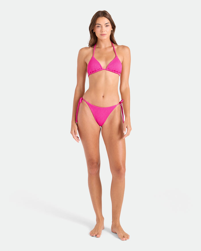 A woman faces forward, wearing the Volcom Women's Pucker Up Triangle Top—a bright pink recycled swimwear bikini with adjustable top and side-tie bottoms—against a plain light background.