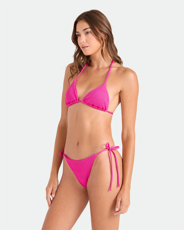 A woman poses against a plain background, wearing the Volcom Women's Pucker Up Triangle Top—a bright pink textured bikini with halter neck ties and side-tie bottoms. The recycled swimwear features an adjustable top for a perfect fit.