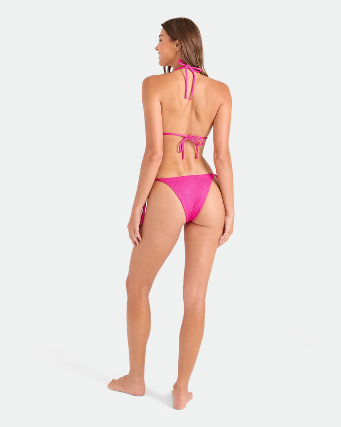 A woman stands barefoot facing away, wearing the Volcom Women's Pucker Up Triangle Top bikini with adjustable halter neck and tie-back, plus matching side-tie bottoms made from recycled swimwear, set against a plain white background.