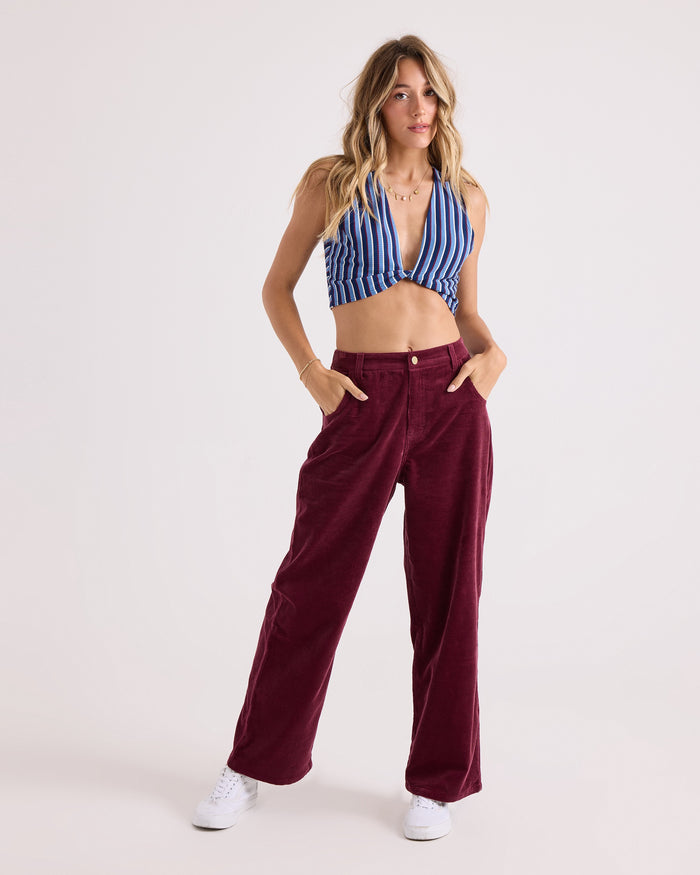 A woman stands with her hands in her pockets, wearing Volcom Women's Relaxed Pull On Cord Pants in maroon, paired with a blue and white striped halter top and white sneakers against a plain light background.