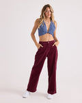 A woman stands with her hands in her pockets, wearing Volcom Women's Relaxed Pull On Cord Pants in maroon, paired with a blue and white striped halter top and white sneakers against a plain light background.