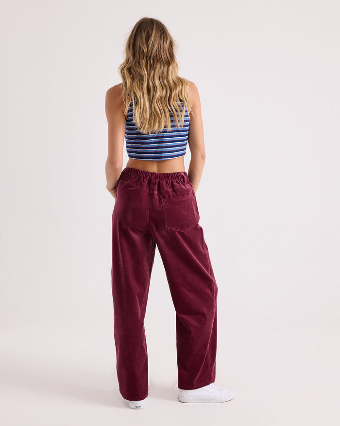 A woman with long wavy hair stands facing away, wearing a blue striped crop top, white sneakers, and Volcom Women's Relaxed Pull On Cord Pants in maroon against a plain light background.