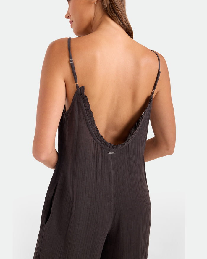 A woman is seen from behind wearing the Volcom Women's Ruffle Up Romper, a loose, dark jumpsuit with thin straps and a deep back. The textured fabric complements her long hair, which drapes over one shoulder.
