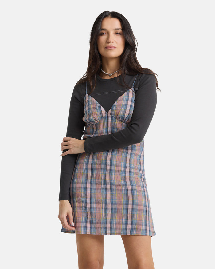 A woman with long dark hair faces forward, wearing the Volcom Women's Sand Burn Dress over a long-sleeve black shirt. She poses with a neutral expression and one arm across her body against a plain white background.