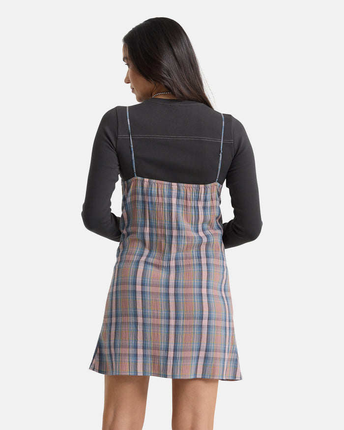 A woman with long dark hair, seen from behind, wears the Volcom Women's Sand Burn Dress—a sleeveless, knee-length plaid dress in blue, pink, and beige—over a black long-sleeve top. The background is plain white.