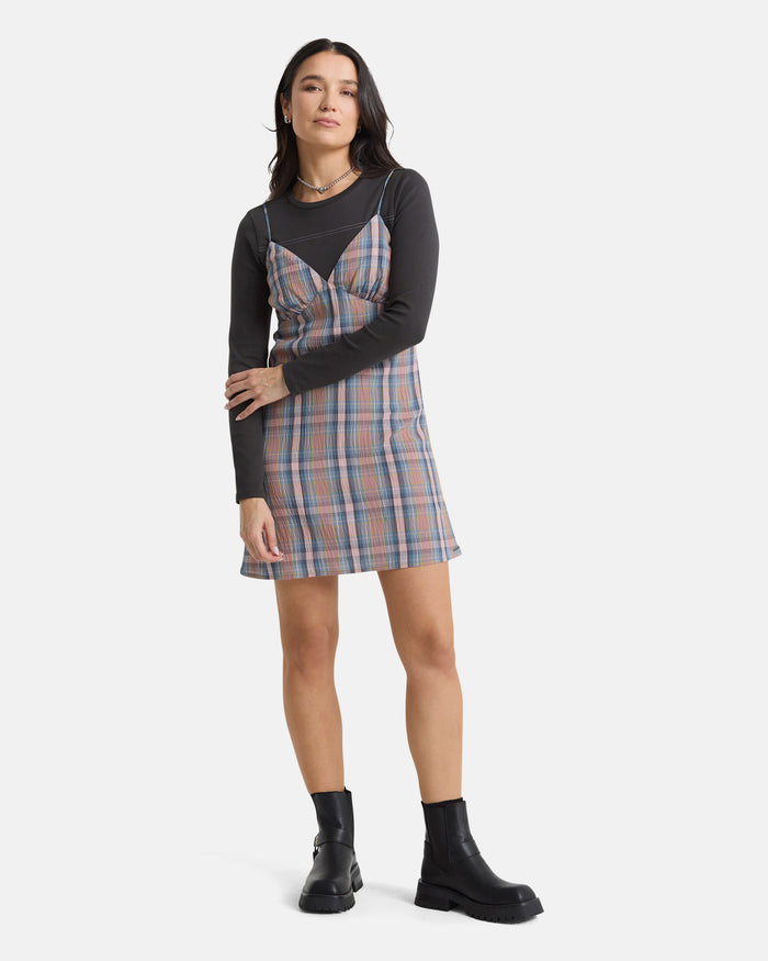 A woman with long dark hair poses confidently against a white background, wearing the Volcom Women's Sand Burn Dress over a black long-sleeve shirt, paired with black ankle boots and layered necklaces.