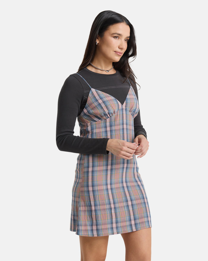 A woman with long dark hair stands against a white background wearing the Volcom Women's Sand Burn Dress in pastel blue, pink, and beige plaid, layered over a fitted black long-sleeve top. She gazes slightly to the side.