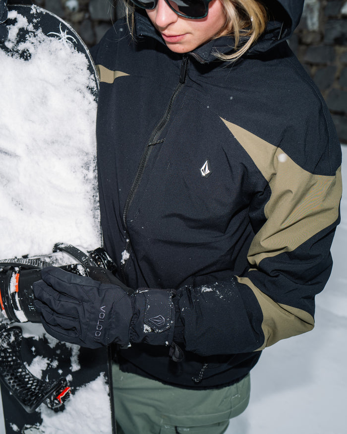 A person wearing the Volcom Women’s Shelter 3D Stretch Jacket in black-and-olive and gloves adjusts snowboard bindings in the snow while wearing sunglasses.