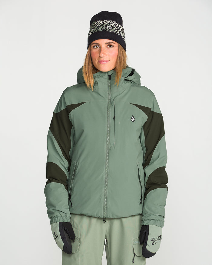 A woman wearing the Volcom Women’s Shelter 3D Stretch Jacket in green and black, featuring waterproof insulation and PFAS-free DWR, paired with matching green pants, black gloves, and a black beanie, stands against a plain white background.