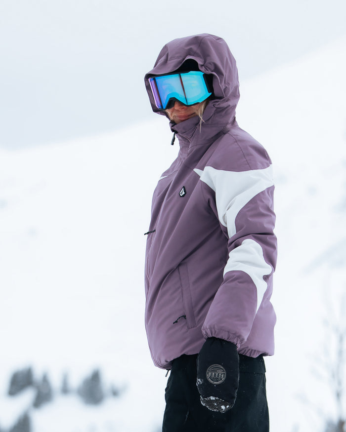 Wearing the Volcom Women’s Shelter 3D Stretch Jacket in purple and white, along with black gloves and reflective ski goggles, a person stands outside amid snowy, mountainous surroundings.