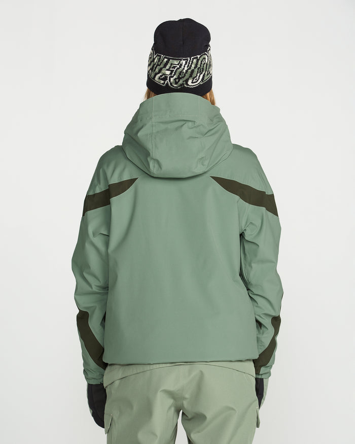 A person wearing the Volcom Women’s Shelter 3D Stretch Jacket in green with PFAS-free DWR, paired with matching green pants, black gloves, and a black beanie featuring white text, stands with their back to the camera against a plain white background.