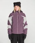 A woman is standing facing forward against a plain white background, smiling slightly, while wearing the Volcom Women’s Shelter 3D Stretch Jacket in purple and gray, paired with black gloves and a black beanie.