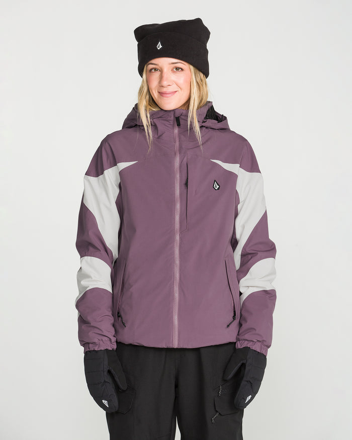 A woman is standing facing forward against a plain white background, smiling slightly, while wearing the Volcom Women’s Shelter 3D Stretch Jacket in purple and gray, paired with black gloves and a black beanie.