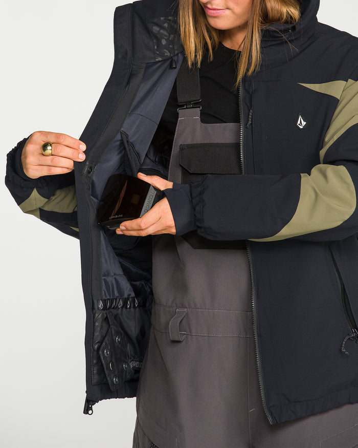 Wearing the Volcom Women’s Shelter 3D Stretch Jacket in black and olive, a person with the jacket unzipped to reveal the inner pocket detail places a smartphone inside, pairing it with gray overalls.