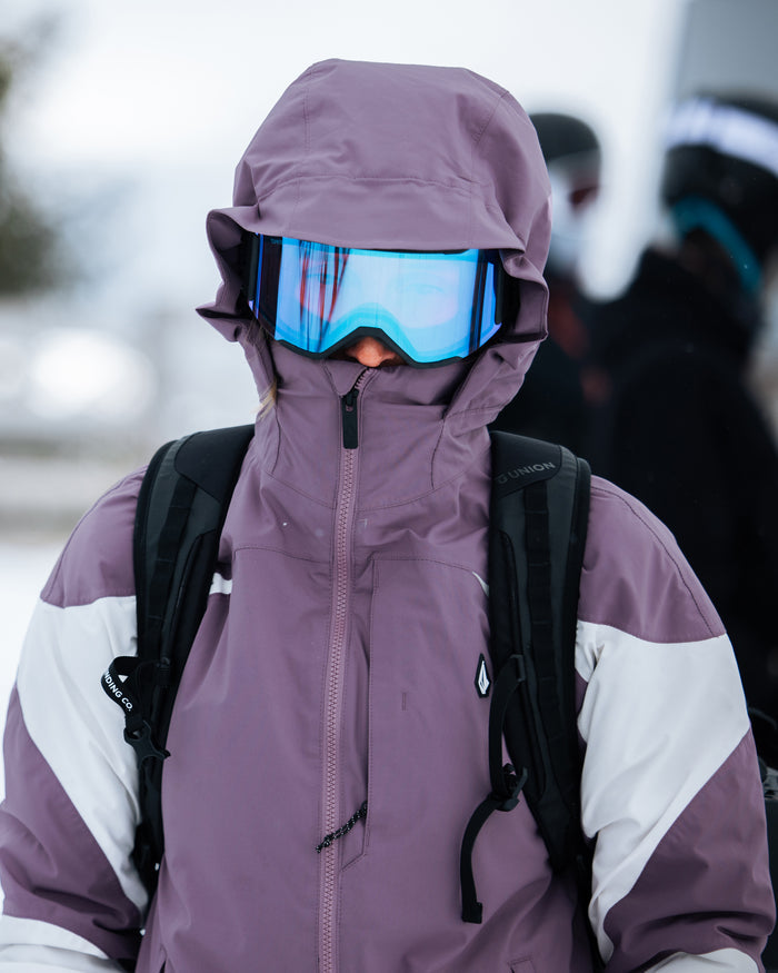 Wearing the Volcom Women’s Shelter 3D Stretch Jacket—with its purple and white colorway, innovative 3D Stretch fabric, and cozy low-loft insulation—a person stands outdoors in snowy conditions, hood up and blue ski goggles on, a backpack slung over their shoulders as blurred figures pass by in the background.