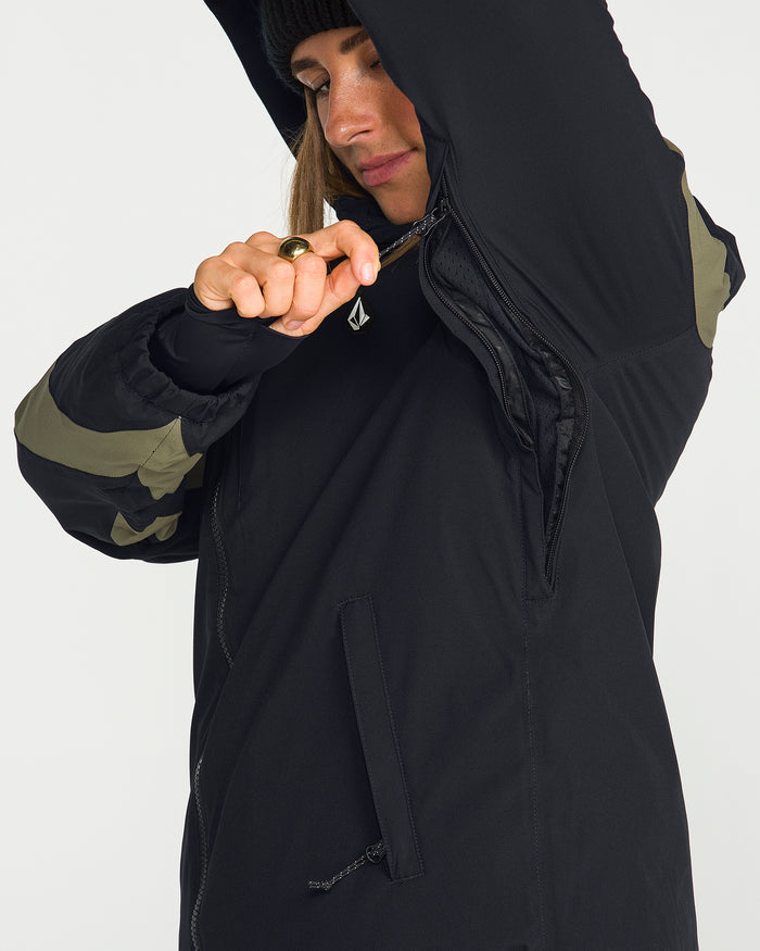 Wearing the Volcom Women’s Shelter 3D Stretch Jacket—a black piece featuring green sleeve accents—a person unzips an underarm zipper to reveal its low-loft insulated interior lining. Partially visible, their face is framed by a black beanie.