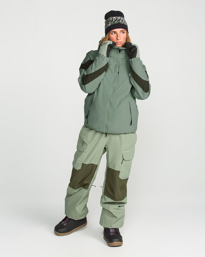 A person is wearing the Volcom Women’s Shelter 3D Stretch Jacket in green with a PFAS-free DWR finish, paired with matching cargo pants featuring dark green patches, black gloves, a black and white beanie, and black boots, all set against a plain white background.