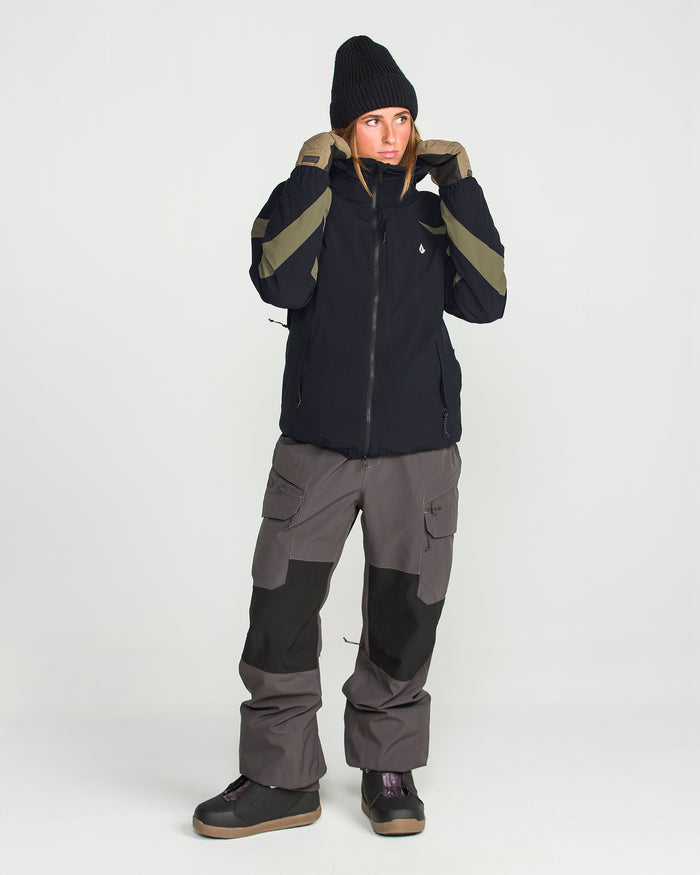 A person is shown wearing the Volcom Women’s Shelter 3D Stretch Jacket—a black winter jacket with green stripes, engineered with advanced 20K/20K defense and low-loft insulation for optimal warmth and protection. They style it with gray cargo pants featuring black knee patches, black boots, and a black beanie, standing against a plain white background.